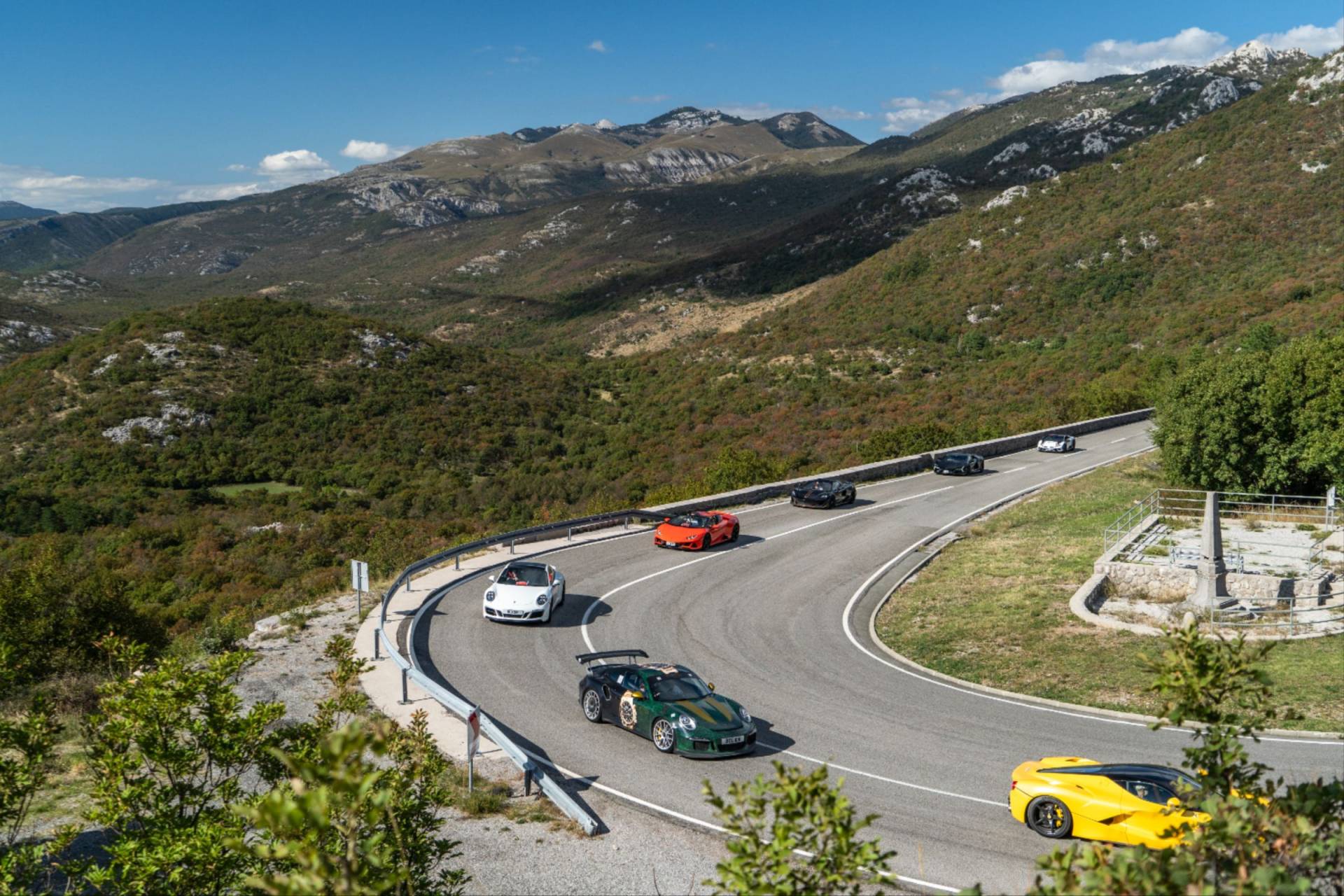 Supercar Driver Club convoy of luxury supercars, including a green Porsche and orange Lamborghini, navigating a scenic mountain road